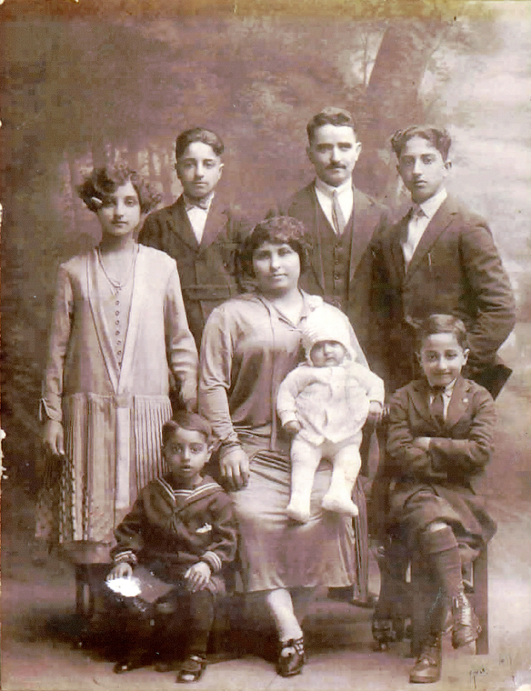 Sepia portrait of family with forest backdrop. Standing from left to right is a daughter, a son, the father, and the oldest son. Sitting from left to right is a toddler son, the mom holding a baby, and another young son. The daughter and mother are wearing light dresses, the father and older sons are wearing dark suits, the toddler son is wearing a dark sailor suit, and the baby is wearing light clothing.