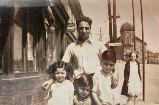 Sepia exterior image taken on sidewalk in front of shops. A man stands in the centre with a girl, and two boys standing in front of him. Another young woman stands in the background right wearing a dress and coat.