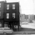 Black and white streetscape. Two attached three-storey townhouses stand on the left with a car parked in front of them. On the right and in the background there is a lot with a parked car and more townhouses. A wire runs across the top half of the image.