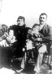 Black and white portrait. Woman wearing a dark dress holds a baby. Man wearing a dark suit holds a toddler. Both the woman and man are sitting on chairs.