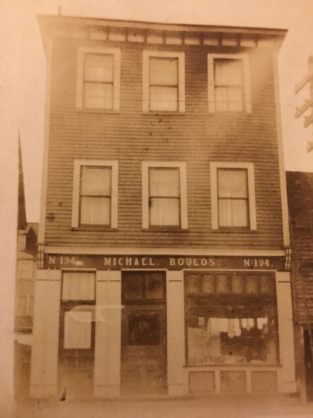 Sepia streetscape. A three storey building is in the centre of the image. It has three windows on the second, and three windows on the third floor. The main floor has two doors, and a large shop window. Above the main floor entrance a sign reads N 194 Michael. Boulos. N 194. The building is attached to a shorter building on the right side, and on the left side you can see a building on the street behind.