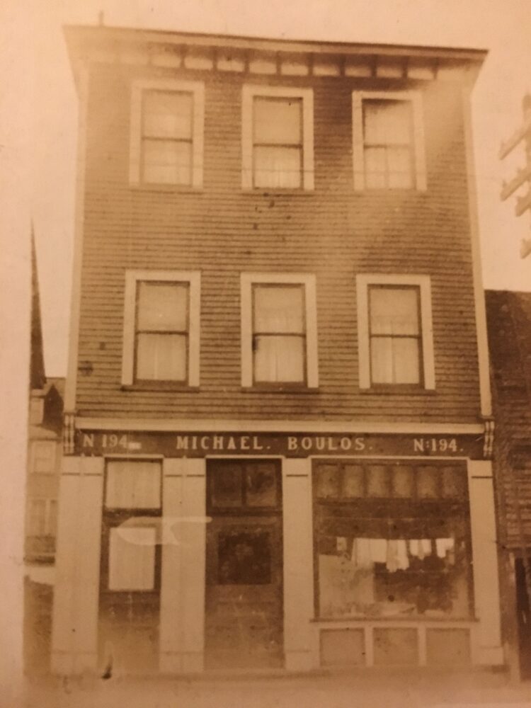 Photo sépia d’un paysage de rue. Un bâtiment de trois étages se trouve au centre de l’image. Il possède trois fenêtres au deuxième et trois fenêtres au troisième étage. L’étage principal comporte deux portes et une grande vitrine de magasin. Au-dessus de l’entrée principale, une enseigne indique N 194 Michael. Boulos. N 194. Le bâtiment est attenant à un autre immeuble moins haut sur le côté droit et, sur le côté gauche, on peut voir un bâtiment dans la rue derrière.