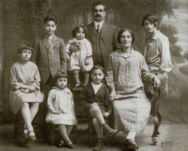 Black and white portrait of family against forest backdrop. Left to right: a seated girl in a light dress and stockings, a standing teenage boy in a suit, a seated young girl in a light dress and stockings, a standing toddler in a light outfit, a man (father) in a dark suit with his right hand on the toddler, a seated young boy in a dark sailor suit, a seated woman (mother) in a light dress, and a young woman in a light outfit. The name Holloway is signed in the bottom right corner.