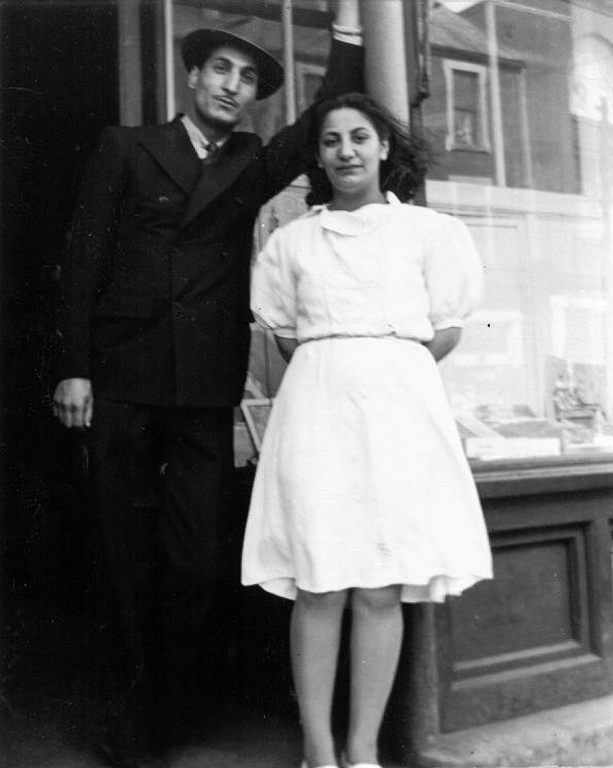 Black and white portrait. A man in a dark suit and fedora with a dark moustache stands on a step with a younger woman with dark hair in a white dress and white shoes. The man is on the left, the woman on the right. The pair stand on the front step in front of a shop. The window behind them reflects the buildings across the street from the store.