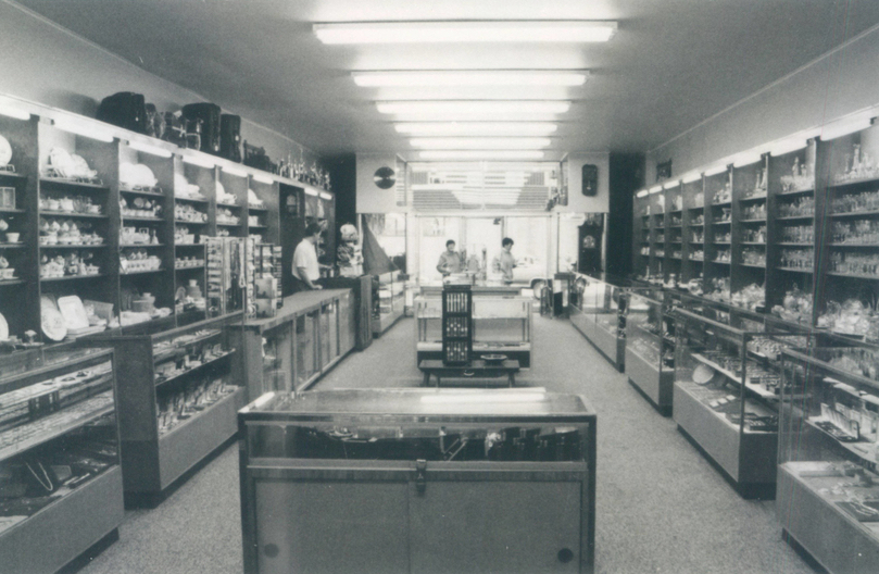 Black and white interior of jewelry store. There are shelves and display cases of jewelry on both sides of the store, two display cases in the middle of the floor, and seven lights in the centre of the ceiling. One man stands behind the counter on the left, and two women stand at the front of the store.