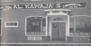 Black and white exterior. Brick building says Ice-cream Parlor; Al. Kawaja’s; Luncheonette Etc. Etc. The two main windows flanking the front door also say Al’s. There is a small roof above the doors.