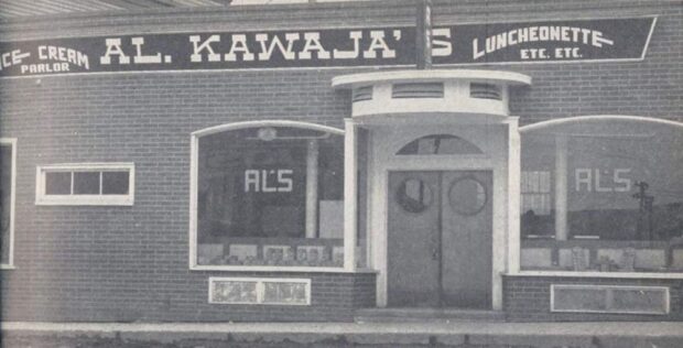 Black and white exterior. Brick building says Ice-cream Parlor; Al. Kawaja’s; Luncheonette Etc. Etc. The two main windows flanking the front door also say Al’s. There is a small roof above the doors.