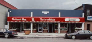 Colour exterior. Brick building with red signage that reads National Shoe; NS; National Shoe; Alteen’s. There is blue paint above the signage. In the middle of the building it says Alteen Bldg 1953. Two cars are parked in front.