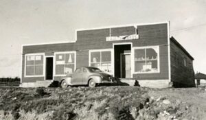 Black and white exterior. Dark building with four large windows, two doors with steps, and a false front. The sign above the main door says Gaultois Bros. There is a car parked in front of the building. Another building can be seen on the right behind this one.