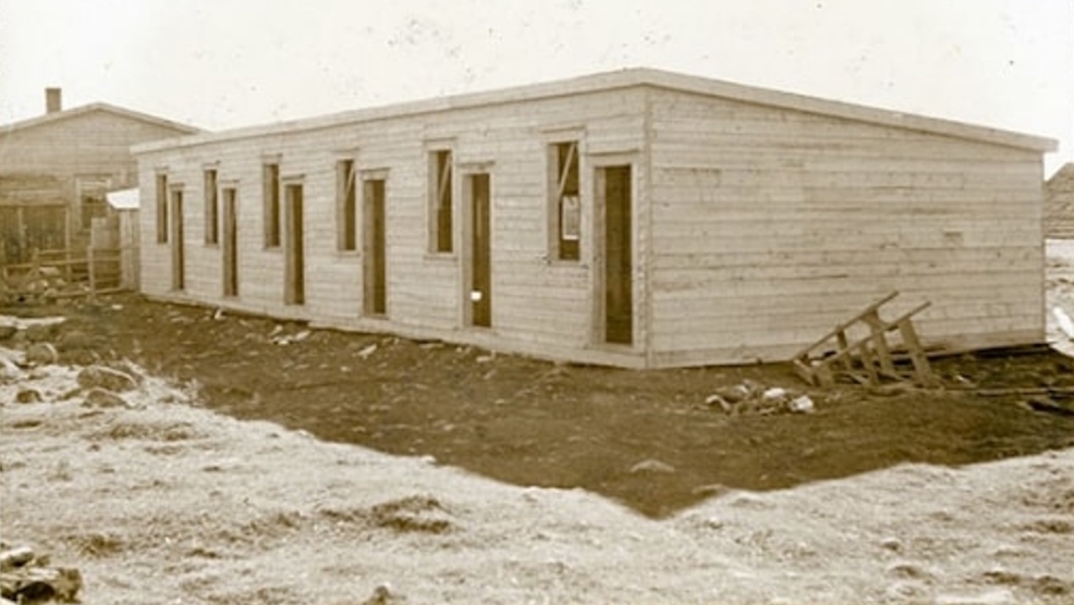 Sepia exterior. Rectangle building under construction. There are six windows and six doors visible. Building appears to be light in colour, and it casts a shadow on the gravel. A step ladder is on the right hand side of the building. There is another building beside this one on the left hand side.