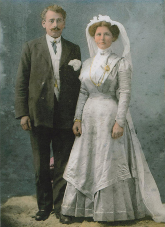 Portrait en couleurs. Un homme portant un costume sombre se tient debout à côté d’une femme portant une robe blanche à manches longues et un voile (qui semble être une robe de mariée). L’arrière-plan est bleu et le couple se tient sur une carpette de couleur brun clair.