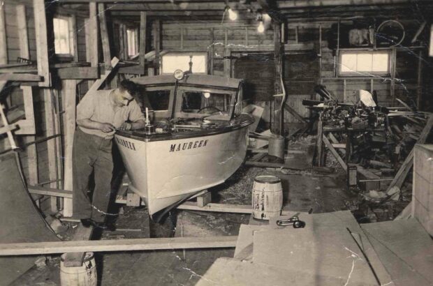 Black and white interior. Man working on a boat inside a wooden shed or outbuilding with five windows. The boat is on the left side of the image and is propped up on wood lumber and barrels. The boat is both light and dark in colour and includes a roof. The words Maureen written twice on either side of the bow of the boat. There are lights along the centre of the ceiling. Miscellaneous wood and tools are on the right side of the image.