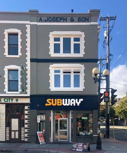 Colour streetscape. Grey building with white windows, and blue accents. Across the top of the building is A. Joseph & Son, and a Subway sign is above the main entrance. Another grey rowhouse is attached on the left. To the right of the building is a street light, a traffic light, and a road that runs perpendicular.