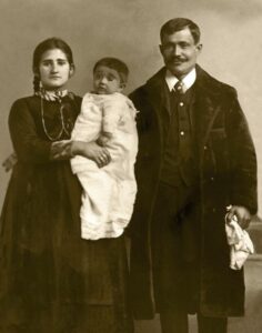 Sepia portrait. Woman wearing a dark dress holds a baby wearing light clothing next to a man wearing a dark suit and jacket.