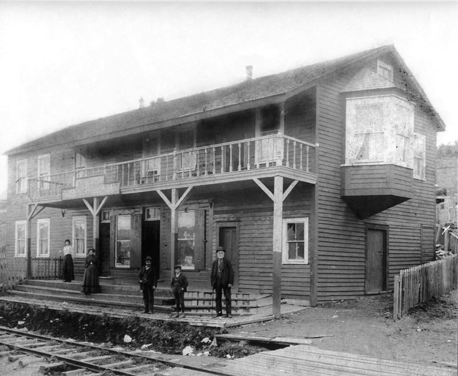 Black and white exterior. Large two storey building in background with balcony in centre and a large bay window on the right. Two women in skirts and dresses, two young boys and an older man in suits stand on the front steps of the building. There is a fence around the building and in front of the steps are railway tracks.