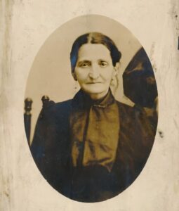 Sepia portrait of woman with dark hair and eyes wearing dark clothes. The woman has her hair up and is sitting in a chair. She is centred in the oval shape of the photo though there is someone next to her.