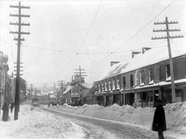 Black and white streetscape. Snow covered street and sidewalks with stone and brick row houses and townhouses on either side as well as utility poles. Approximately twelve people are on the sidewalk. There is one man on the street on the right side in the foreground of the photo. There is another man on the right side of the street leading a horse. A snow plow is clearing the left side of the street.