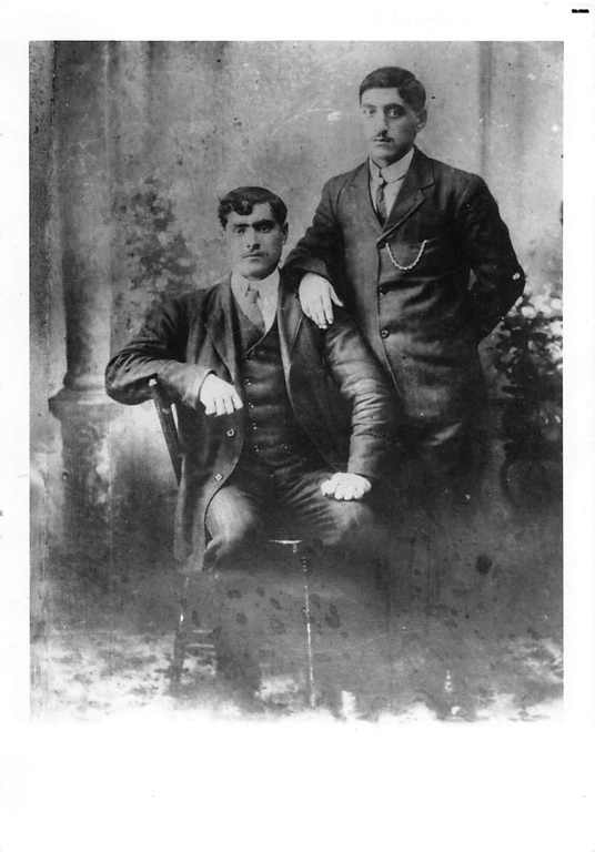 Black and white portrait. Two men in suits side by side. The man on the left is sitting on a chair while the man on the right is standing. The backdrop features a column, and several plants.