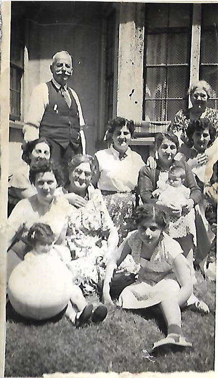 Black and white portrait. 11 people sitting outside the front of a building. There are six women sitting and kneeling, one woman standing at the back right, and one man standing at the back left. One of the women on the right is holding a baby and there is a little girl and a toddler sitting in the front. The women and girls are all wearing dresses or skirts and shirts, and the man is wearing a dress shirt, vest, tie, and pants.