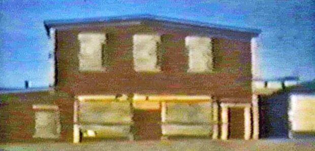 Colour still image taken from a video. Two storey house with six windows, one door, and an addition on the left side. To the right of the house is a small building.