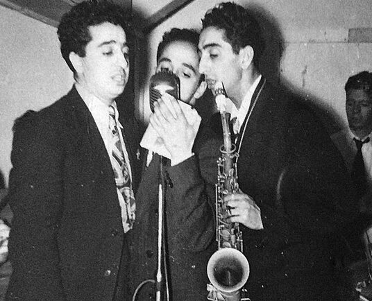 Black and white portrait of three men. The men all have dark hair and are all wearing dark suits and singing into a microphone. The one on the right holds a saxophone. In the background behind the man on the right is another man wearing a white dress shirt and dark tie.