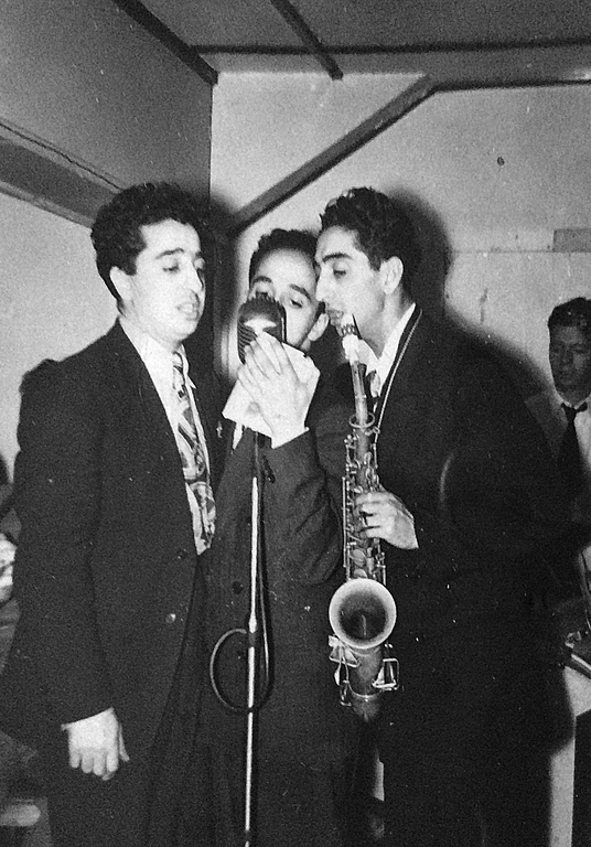 Black and white portrait of three men. The men all have dark hair and are all wearing dark suits and singing into a microphone. The one on the right holds a saxophone. In the background behind the man on the right is another man wearing a white dress shirt and dark tie.