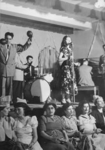 Photo d’intérieur en noir et blanc. Groupe de personnes faisant partie d’un orchestre et se produisant sur une scène. Le groupe est composé de six personnes, cinq hommes et une femme. La femme chante tandis que les hommes jouent de différents instruments (piano, saxophone, maracas, batterie, etc.). En bas de la scène, six femmes sont assises, le dos tourné à la scène.