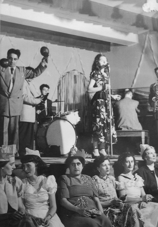 Black and white interior. Group of people in a band performing on a stage. The band consists of six people, five men, and one woman. The woman sings while the men play a variety of different instruments (piano, saxophone, maracas, a drum set, etc.) Below the stage are six women sitting down with their backs turned from the stage.