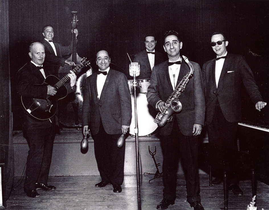 Black and white portrait. Band that consists of six men dressed in suits each holding a different instrument (from left to right guitar: cello, maracas, drum kit, saxophone, and piano). There is a microphone in the very centre of the photo, and the cello player and drummer are standing and sitting on the stage above the four other men standing in front.