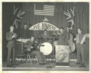 Black and white interior. Band called The Jive Bombers has three men on a stage in the back and four men in the front. All seven men are wearing suits or military uniforms and playing an instrument. From left to right clarinet, vibraphone, guitar, drum kit, trumpet, cello, and accordion. There are three flags hung on the curtains behind and above the men, two Union Jack flag, and one American flag in the centre. Below the American flag there is a sign for the Jive Bombers.