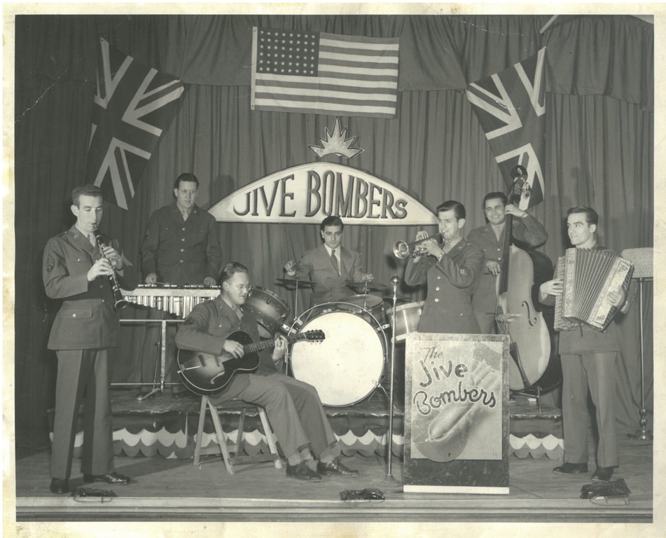 Black and white interior. Band called The Jive Bombers has three men on a stage in the back and four men in the front. All seven men are wearing suits or military uniforms and playing an instrument. From left to right clarinet, vibraphone, guitar, drum kit, trumpet, cello, and accordion. There are three flags hung on the curtains behind and above the men, two Union Jack flag, and one American flag in the centre. Below the American flag there is a sign for the Jive Bombers.