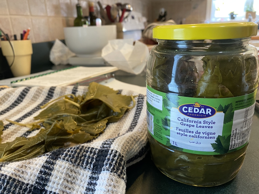 Closeup colour photo of kitchen. A bottle of California style grape leaves on the right is the focus of the image. To the left of the bottle you can see a plate covered in a dish towel with some grape leaves drying on it. In the background you can see a cookbook and kitchen utensils on a counter, and a stove.