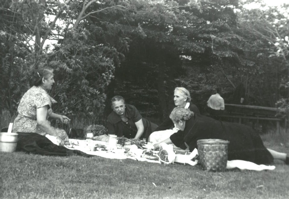 Black and white exterior. Four older women having a picnic in a park. The women are all wearing dresses and two are sitting while two are lying down on the picnic blanket. There are a variety of different foods and two baskets on top of a light coloured blanket. There are trees in the background as well as a railed fence, and a man in the background right.