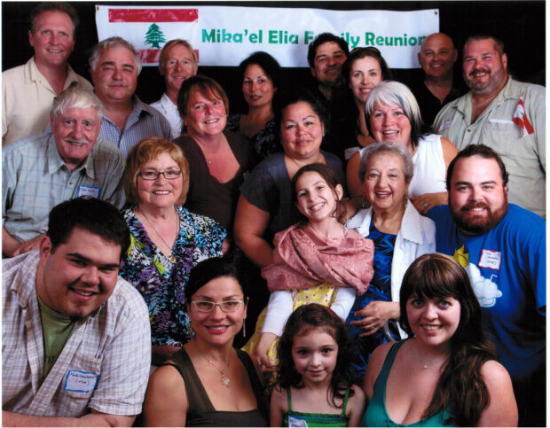 Colour portrait. Group of 20 people, nine men, nine women, and two young girls posed in front of a sign with the Lebanese flag in the left corner (red and white flag with green tree in centre), to the right of the flag is green text that says Mika’el Elia Family Reunion.