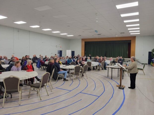 Colour interior. Large crowd of approximately 70 people who are all sitting around 10 tables all watching a woman doing a presentation. The group is a mix of men and women of all ages. The woman presenting is standing at the front of the room at a podium. There is blue tape on the floor that making a circular pattern near the podium. In the background of the photo there are three blue doors, and in between two of the doors there is a big stage with green curtains.