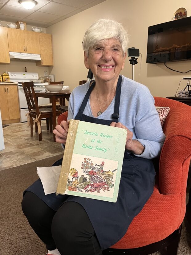 A woman with short hair holds a cookbook that says Favorite Recipes of the Basha Family. She is sitting on a red chair wearing a light blue long sleeve shirt, a dark blue apron and black pants.