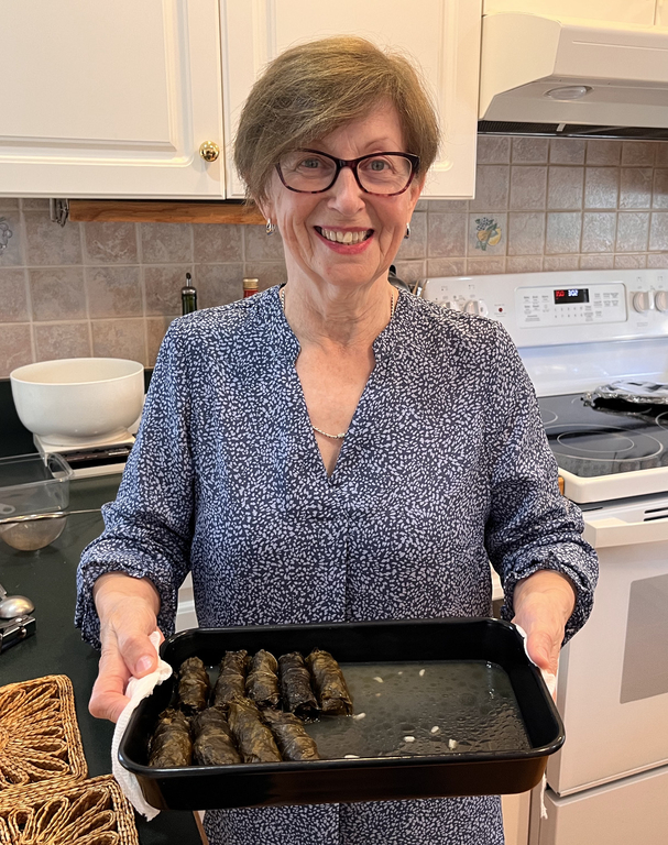 A woman with short hair and wearing glasses, and a blue and white blouse holds a pan of nine grape leaves. She is standing in a kitchen.