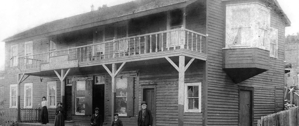 Black and white exterior. Large two storey building in background with balcony in centre and a large bay window on the right. Two women in skirts and dresses, two young boys and an older man in suits stand on the front steps of the building. There is a fence around the building and in front of the steps are railway tracks.