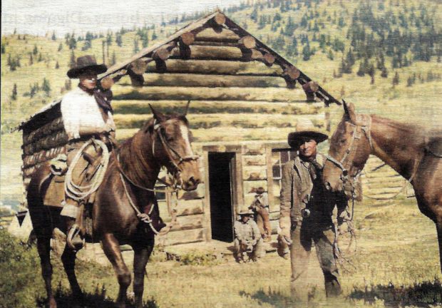 Two cowboys with horses before a log cabin