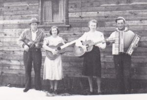 A group of four musicians stand beside a rustic wooden building. Two women have guitars. One man is holding a fiddle and the other man has an accordion.