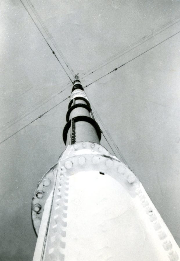 A radio tower, as seen when looking from its base straight up to the top. A series of support wires extend outward in four directions.