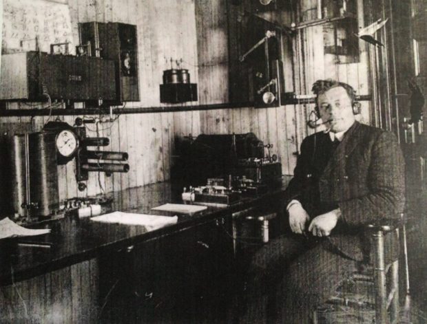 A man in a suit sits facing the camera beside a desk topped by wireless equipment and neat piles of paper. He wears a set of wired headphones and smokes a pipe.