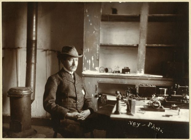 A man in a checked jacket and hat sits at a table topped by wireless equipment. Behind him are bare cupboards with no doors, and a metal stove and stovepipe.