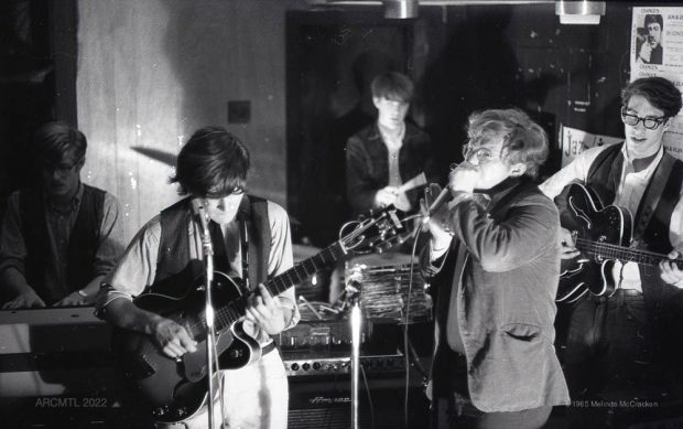 Black and white photograph of five musicians performing in the corner of a wood-paneled room. Two guitar players and a harmonica player are seen in the foreground, a drummer and organ player are seen in the background.