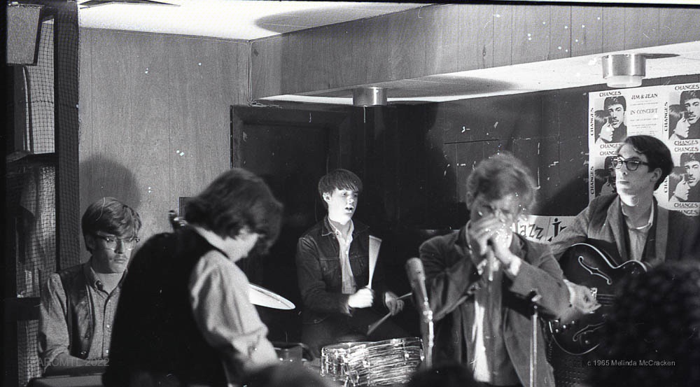 Black and white photograph of five musicians performing in the corner of a wood-paneled room.