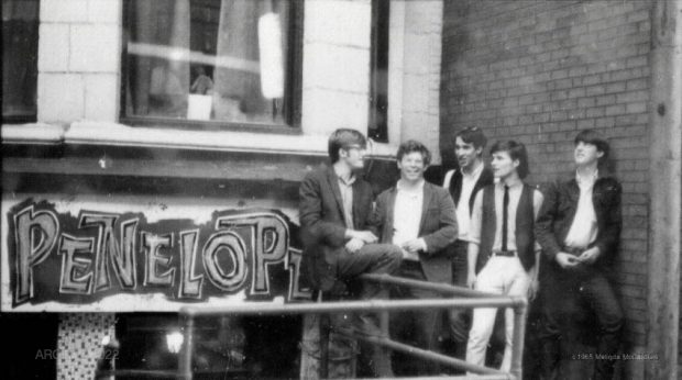 Black and white photograph of five young members of the group Sidetrack standing casually outdoors in front of a brick wall and a sign that reads Penelope.