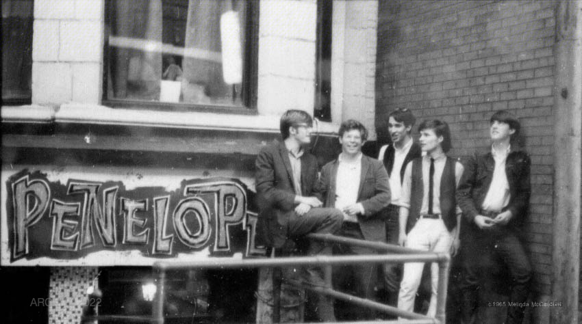 Photographie en noir et blanc de cinq jeunes membres du groupe Sidetrack se tenant nonchalamment en plein air devant un mur de briques et une pancarte indiquant Pénélope.