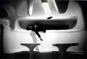 A black and white photograph of a man with glasses lying on a molded curved white alcove with one arm hanging down. Molded white tables can be seen in the foreground. A white neon light coming from a tube-like form is hanging from the ceiling into the centre of the image.