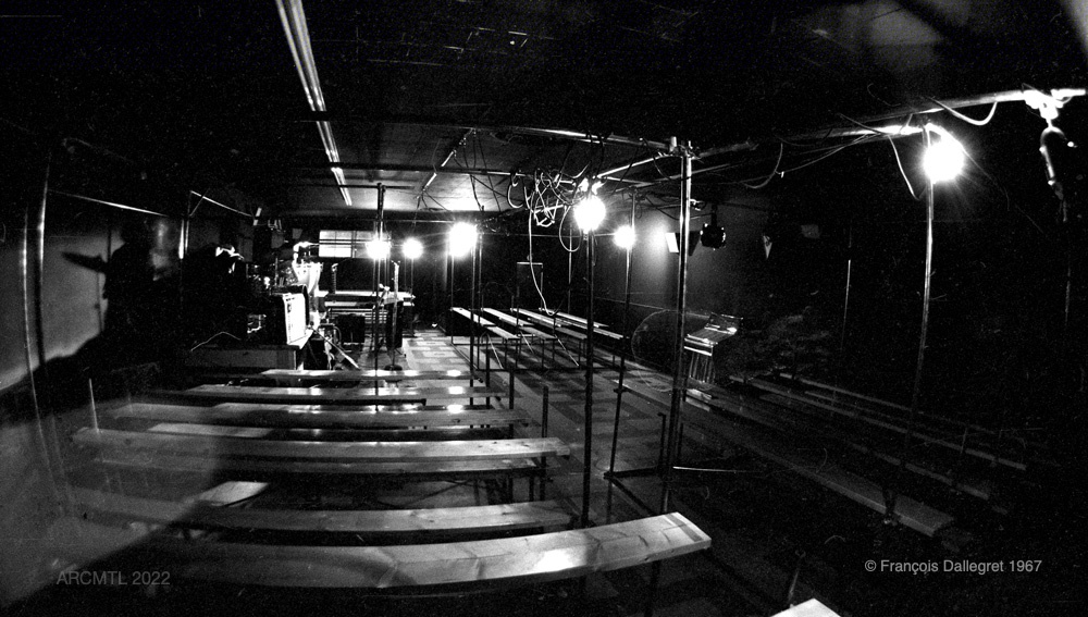 A high-contrast black and white wide-angle photograph of the completely empty interior of The New Penelope, showing rows of benches and bright lighting fixtures hanging from scaffolding overhead.