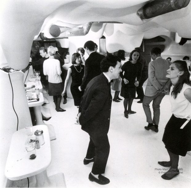 A black and white photograph of a dozen people dancing in a brightly-lit room with white bulbous moulded curved walls and curved ceiling. White tables are placed along the wall on the left side of the image.